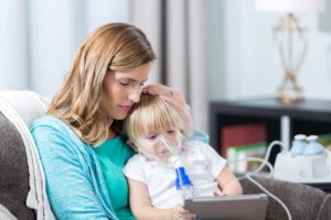 Toddler boy with nebulizer sitting on mom's lap playing with tablet