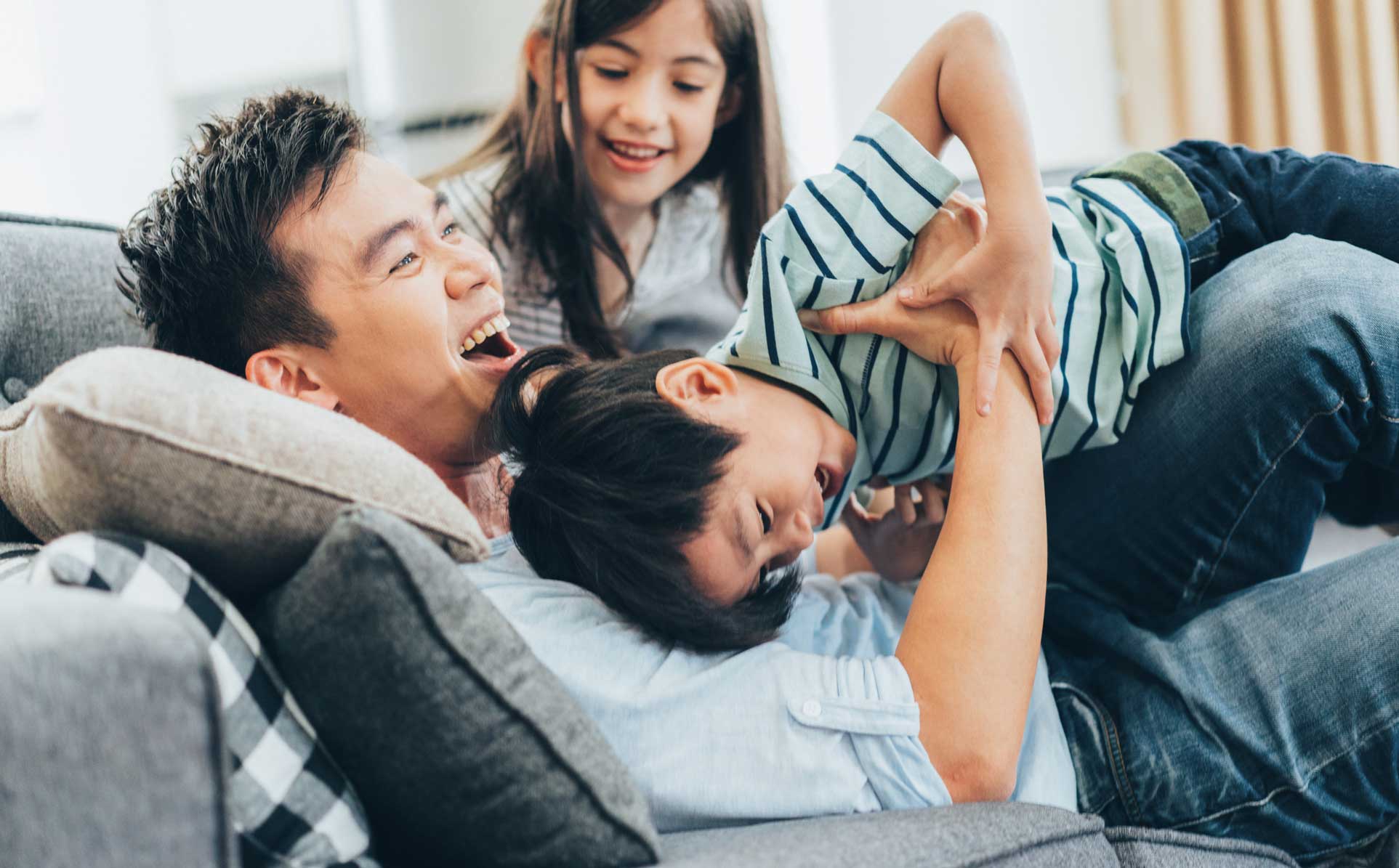 Dad tickling son on couch, daughter in background smiling